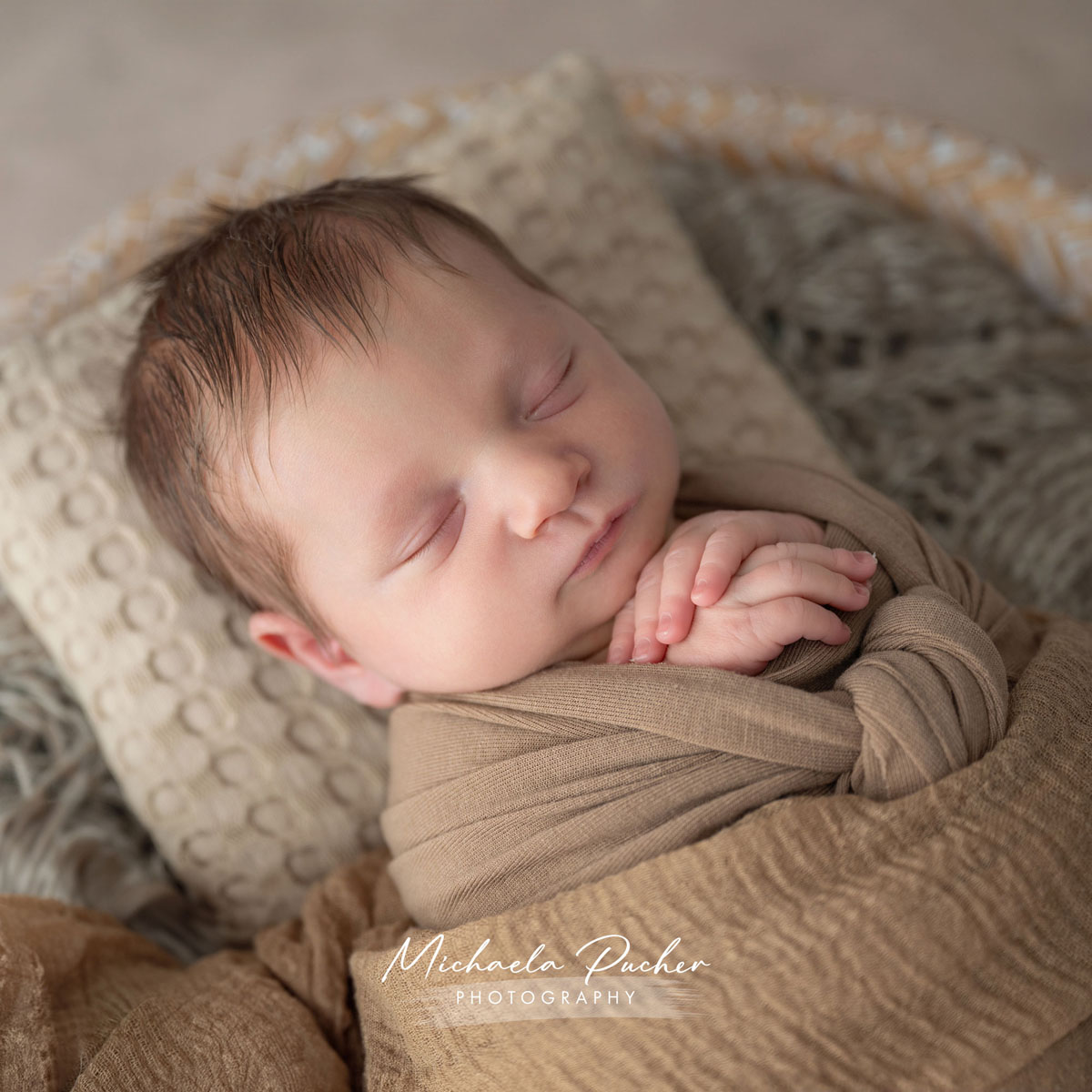 Schlafendes Neugeborenes, eng eingewickelt in braune Tücher, liegt mit gefalteten Händen auf einem strukturierten Kissen in warmen Naturtönen.